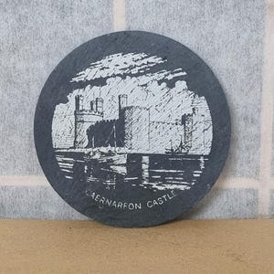 Könnte beinhalten: Ein schwarzer Schieferuntersetzer mit einer weißen Gravur von Caernarfon Castle. Die Burg ist mit einem Wassergraben und Wasser dargestellt, das sie umgibt. Der Text "Caernarfon Castle" steht unter dem Bild.