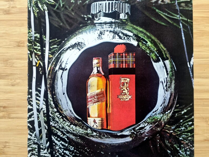 Puede incluir: Un anuncio con una botella de whisky Johnnie Walker y una caja de regalo roja con un patr&oacute;n de cuadros, dentro de una gran bola de Navidad plateada. La caja de regalo tiene un escudo y el texto "150 1820-1970". El fondo incluye ramas de hoja perenne.