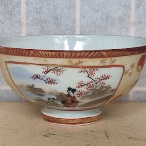 May include: A white ceramic bowl with a gold rim and a painted scene of a woman in a red kimono standing under a cherry blossom tree. The bowl has a small foot.