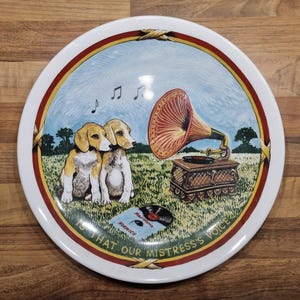 May include: A white ceramic plate with a red, yellow, and green border. The center of the plate features a colorful illustration of two dogs listening to a vintage gramophone playing a record. The text on the plate reads "Is that our mistress's voice".