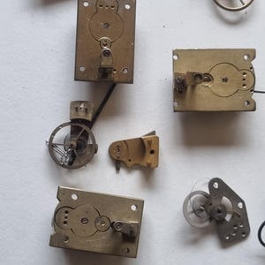 May include: A collection of vintage watch parts, including brass plates, gears, and springs. The components are in various shapes and sizes, with intricate details visible. The image showcases the mechanical elements of timekeeping devices.