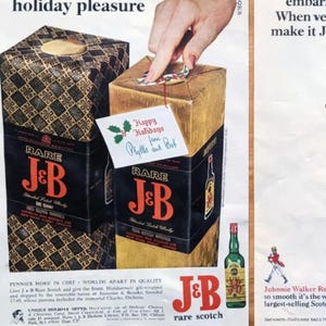 May include: Vintage advertisement featuring two gift boxes of J&B Rare Scotch whisky. The boxes are gold and black, with the brand name prominently displayed. A hand is shown opening one box, with a holiday greeting card attached. A green bottle of whisky is also visible.