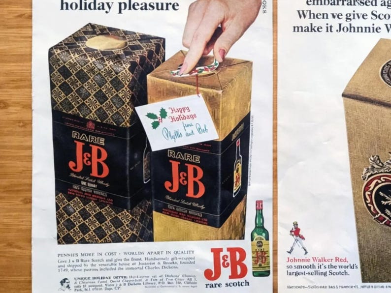 May include: Vintage advertisement featuring two gift boxes of J&B Rare Scotch whisky. The boxes are gold and black, with the brand name prominently displayed. A hand is shown opening one box, with a holiday greeting card attached. A green bottle of whisky is also visible.