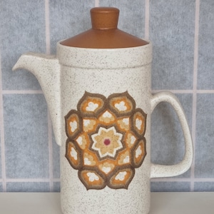 Könnte beinhalten: Eine weiße Kaffeekanne aus Keramik mit braunem Deckel und einem geometrischen Muster in Braun-, Orange- und Weißtönen. Die Kanne hat einen Griff und einen Ausguss.