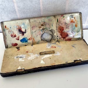 May include: An open, vintage watercolor paint box with a black frame and a cream-colored interior. The paint box is divided into three sections, each with dried paint in various colors. A small, rectangular metal plate is visible on the inside.