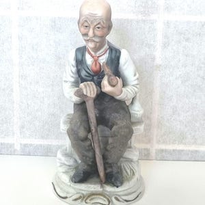 Puede incluir: Una figurita de porcelana de un hombre mayor con bigote y pipa, sentado en un banco con un bastón. Lleva una camisa blanca, un chaleco negro y pantalones marrones. La figurita está pintada en colores realistas.