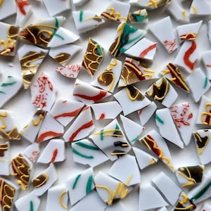 Könnte beinhalten: Nahaufnahme von zerbrochenen weißen Keramikscherben, die verstreut sind. Die Fragmente sind mit roten, grünen und goldenen Mustern verziert, einige zeigen asiatische Kalligraphie. Die Anordnung erzeugt einen Mosaikeffekt.