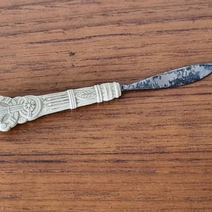 May include: An antique butter knife with a decorative ivory-colored handle and a tarnished metal blade. The handle features an ornate design with floral and geometric patterns. The knife is set against a wooden surface.