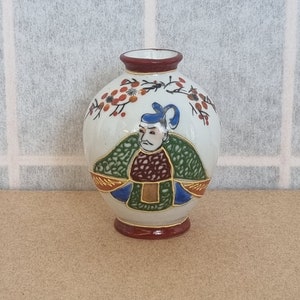 Puede incluir: Pequeño jarrón de cerámica blanca con cuerpo redondeado y cuello estrecho. Presenta una ilustración pintada de una figura con vestimenta tradicional, con sombrero azul y túnica verde. Adornos florales rojos y naranjas decoran la parte superior.