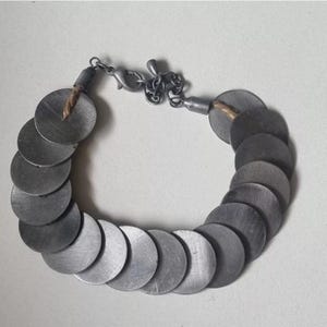 May include: A silver-toned bracelet composed of numerous circular metal discs. The discs are linked together, forming a flexible chain. The bracelet features a clasp closure and a small chain for adjusting the length. The overall design is simple and modern.