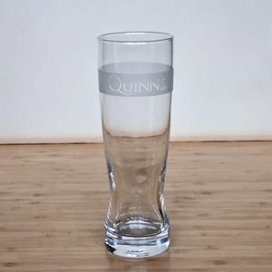 Puede incluir: Un vaso de cerveza alto y transparente con una base acampanada y un cuerpo cilíndrico. El vaso tiene una banda gris con el nombre "Quinn's" impreso. El vaso está vacío y sobre una superficie de madera.
