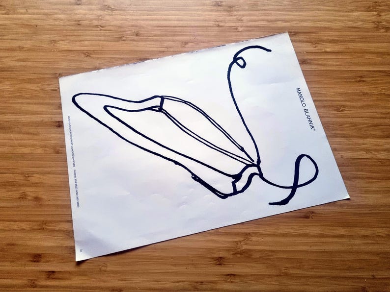 May include: A white paper with a black line drawing of a shoe, with the text "MANOLO BLAHNIK" in black. The paper is on a wooden surface. The shoe design has a curved shape with a long, looping line.