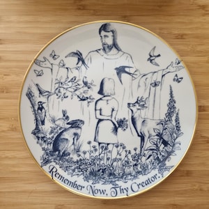May include: Decorative white plate with a gold rim featuring a blue and white illustration. The design includes a figure with outstretched arms, a girl, deer, birds, butterflies, and the text "Remember Now, Thy Creator."