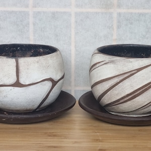 May include: Two white ceramic planters with brown geometric designs. The planters are round and have a slight lip around the top. They are sitting on brown saucer-shaped bases.
