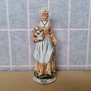 Könnte beinhalten: Eine Keramikfigur einer Frau in einer weißen Schürze und einem braunen Kleid, die einen Blumenstrauß hält. Sie lächelt und trägt eine weiße Mütze auf dem Kopf.