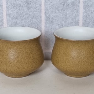May include: Two brown ceramic mugs with white interiors. The mugs have a speckled texture.