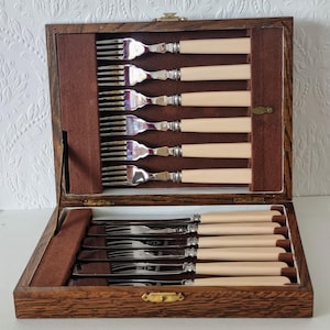 May include: A wooden cutlery set with a hinged lid, featuring forks and knives with cream-colored handles and silver tines and blades. The set is displayed in a dark brown wooden box with a brass clasp.