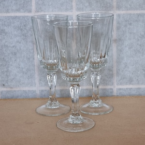 May include: Three clear, stemmed glasses with a classic design. Each glass features a detailed stem and a flared bowl with vertical etched lines. The glasses are arranged on a light brown surface against a backdrop of a white and gray grid.