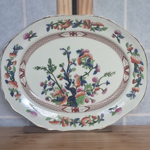 Pode incluir: Um prato oval para servir com fundo branco e um desenho floral em tons de azul, verde, laranja e vermelho. O desenho floral apresenta uma árvore central com ramos e folhas, rodeada por flores e folhas menores. O prato tem uma borda dourada e uma borda festonada.