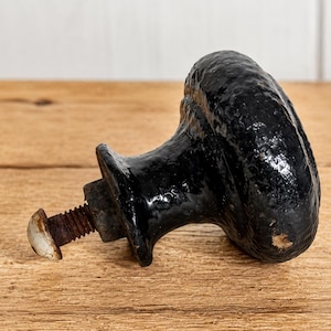 May include: A black, antique-style cabinet knob with a textured, oval-shaped handle. The knob is attached to a rusty bolt with a white head. The hardware is set against a wooden surface.