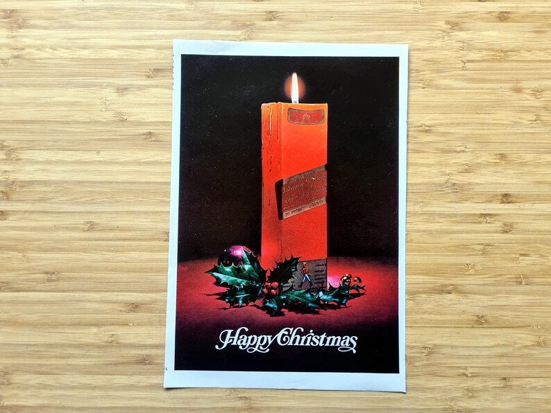 May include: A vintage Christmas card showing a lit orange candle with a label. The candle is on a red surface with holly and a purple bauble. The words "Happy Christmas" are written in white script.