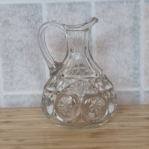 May include: A clear glass pitcher with a textured surface and a round body. The pitcher has a handle and a spout.