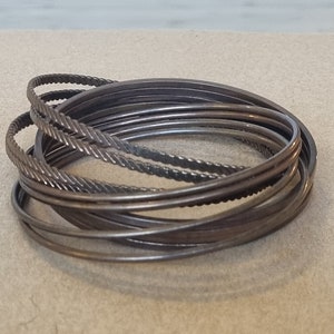 Può includere: Una collezione di braccialetti color argento. Alcuni braccialetti hanno una finitura liscia e lucida, mentre altri presentano disegni strutturati come motivi intrecciati o attorcigliati. I braccialetti sono impilati, creando un look a strati.