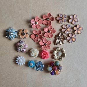 Pode incluir: Conjunto de botões e enfeites em forma de flor de várias cores, incluindo rosa, pêssego, azul, branco e dourado. Alguns botões têm strass ou contas.