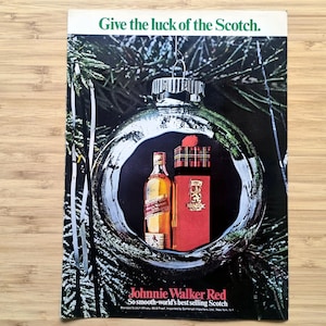 Vintage 1960s Johnnie Walker Red Christmas advertisement showing a bottle and boxed gift set displayed inside a reflective silver ornament hanging on a pine tree. The headline reads “Give the luck of the Scotch.” The full printed page is laid flat on a wood surface, showing its colours and rectangular shape.