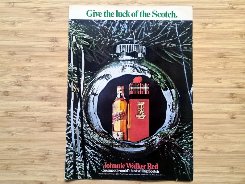 Vintage 1960s Johnnie Walker Red Christmas advertisement showing a bottle and boxed gift set displayed inside a reflective silver ornament hanging on a pine tree. The headline reads “Give the luck of the Scotch.” The full printed page is laid flat on a wood surface, showing its colours and rectangular shape.