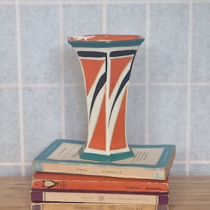 May include: A stack of vintage paperback books with colourful covers. The top book has the title "Walden" by Thoreau. The books are stacked on a wooden surface. A geometric orange, black and white ceramic vase sits on top of the books.