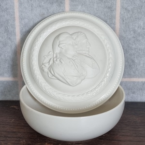 Könnte beinhalten: Weiße Keramik-Schmuckdose mit einem Relief von zwei Personenköpfen. Die Dose hat einen runden Deckel und eine schalenförmige Basis.