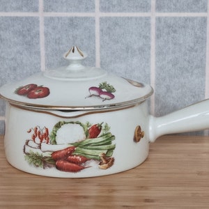Może przedstawiać: Biała ceramiczna naczynie do zapiekania z pokrywką. Naczynie ma złotą obwódkę i kolorowy wzór warzyw. Pokrywka ma gałkę w kształcie kwiatu.