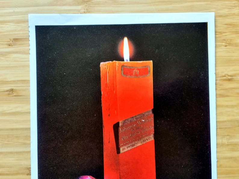 May include: A lit, rectangular red candle with a label that reads "Johnnie Walker Old Scotch Whisky". The candle is set against a black background. The flame is bright and yellow.