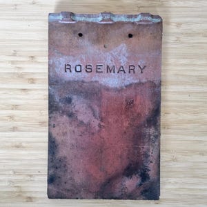 Può includere: Una tegola rettangolare in terracotta usurata con la parola "ROSEMARY" in rilievo al centro. La tegola ha un colore marrone-rossastro screziato con aree più scure e due piccoli fori vicino alla parte superiore. Il bordo superiore ha un aspetto strutturato e irregolare.