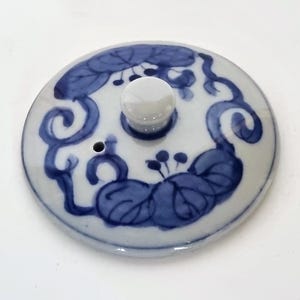 Puede incluir: Tapa redonda de cerámica con base blanca y diseño floral azul. La tapa presenta un pequeño pomo blanco en el centro y un pequeño agujero. El diseño azul incluye hojas y espirales.