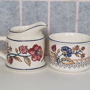 May include: A cream-coloured ceramic creamer and sugar bowl set with a floral design. The creamer has a handle and a spout. The sugar bowl is round and has a lid. The floral design features red, yellow, blue, and green flowers.