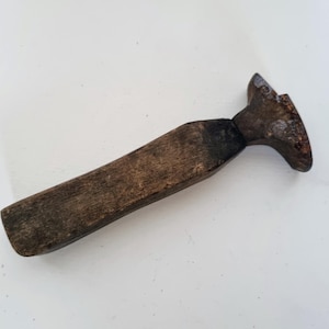 Puede incluir: Una herramienta de madera envejecida, con un mango largo y rectangular y una cabeza curvada en forma de paleta. La madera tiene un aspecto oscuro y envejecido, lo que sugiere que es un objeto antiguo. La herramienta probablemente se utiliza para dar forma o alisar materiales.
