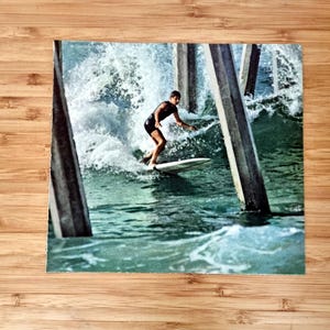 Może przedstawiać: Zdjęcie w stylu vintage przedstawia surfera na fali. Surfer, ubrany na czarno, balansuje na białej desce surfingowej w wzburzonej wodzie. Scena jest otoczona przez zwietrzałe drewniane filary, co wywołuje nostalgię i emocje związane z surfingiem.