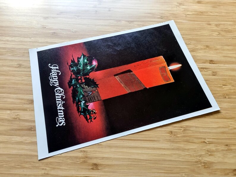 May include: A Christmas card with a festive design. The card features a red candle with a lit flame, holly with red berries, and the words "Happy Christmas" in white script. The background transitions from red to black.