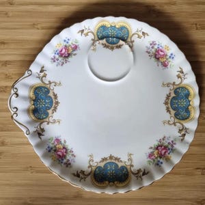 Puede incluir: Plato de cerámica blanco con borde festoneado y una hendidura central. El plato presenta ramos florales y adornos decorativos azules y dorados. Un borde dorado bordea el borde del plato.