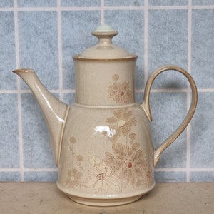 May include: A beige ceramic teapot with a floral design. The teapot has a lid with a light green knob and a handle on the side. The teapot is decorated with brown and cream colored flowers.