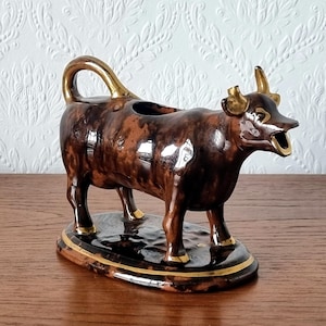 May include: A brown ceramic cow creamer with gold accents. The creamer is in the shape of a standing cow with a handle and spout. The horns, handle, and base are gold. The creamer is on an oval base.
