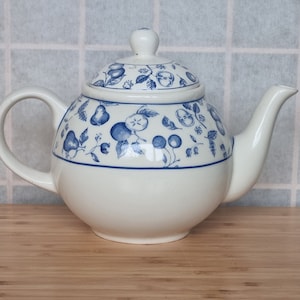 Puede incluir: Una tetera de cerámica blanca con tapa, asa y boquilla. La tetera está decorada con motivos azules de frutas y hojas. Una franja azul rodea el cuerpo de la tetera. La tetera está sobre una superficie de madera.