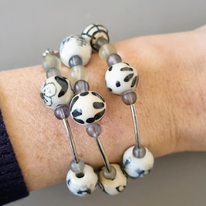 May include: A triple-strand bracelet with white ceramic beads featuring black and white patterns. The beads are strung on silver wire with gray spacer beads.