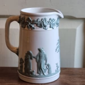 May include: A white ceramic pitcher with a green floral design and a raised relief scene of people and a dog. The pitcher has a handle and a spout.