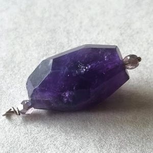 Puede incluir: Un colgante de piedra preciosa de amatista púrpura oscuro facetada con pequeñas cuentas redondas púrpuras en cada extremo. La piedra preciosa tiene forma de hexágono alargado. El colgante está sobre un fondo gris claro.