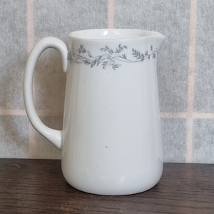 Op de afbeelding: Witte keramische kan met een gebogen handvat en een grijs bloemmotief rond de rand. De kan heeft een klassieke vorm en is geschikt voor het serveren van dranken. De kan staat op een houten oppervlak.