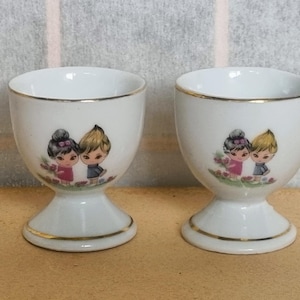Puede incluir: Dos hueveras de cerámica blanca con ribete dorado. Cada taza presenta una imagen pintada de un niño y una niña. Las hueveras tienen una base de pedestal y son adecuadas para servir huevos.