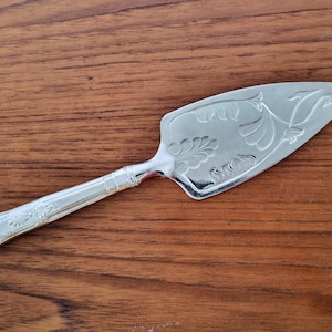 Op de afbeelding: Zilveren taartschep met een sierlijk bloemen- en bladerpatroon. De schep heeft een driehoekig blad en een lange handgreep. De handgreep en het blad zijn versierd met gedetailleerde gravures. Het wordt gepresenteerd op een houten oppervlak.